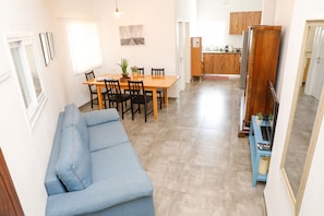 A modern living room with a blue sofa, a wooden dining table with chairs, and a kitchen area with wooden cabinets and a refrigerator.