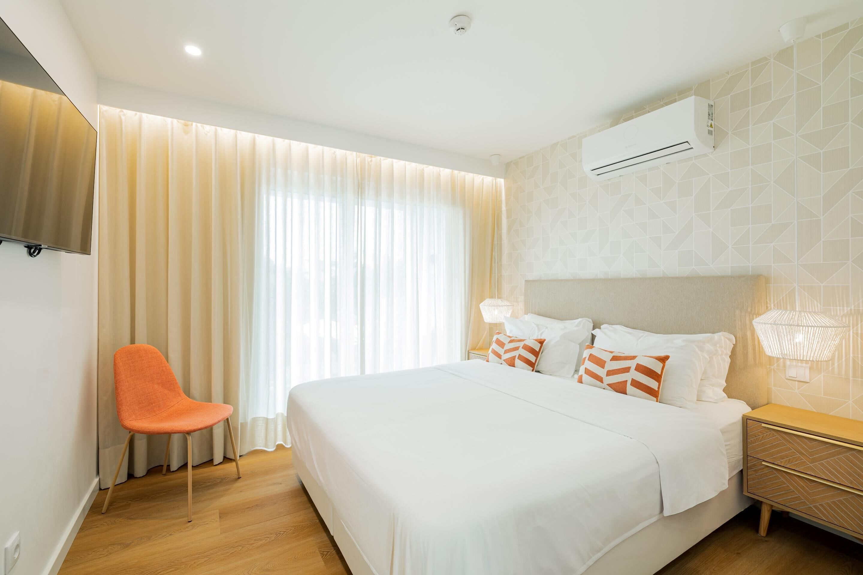 A hotel room with a large bed, a chair, a nightstand, an air conditioning unit, and a window with curtains.