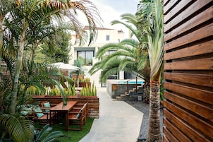 Un patio con muebles de madera, palmeras y un sendero de concreto que lleva a un edificio con puerta de vidrio.