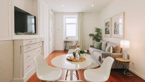 A modern living room with a round white table, two white chairs, a flat-screen TV mounted on a cabinet, a sofa, a small side table with a lamp, and framed artwork on the wall.