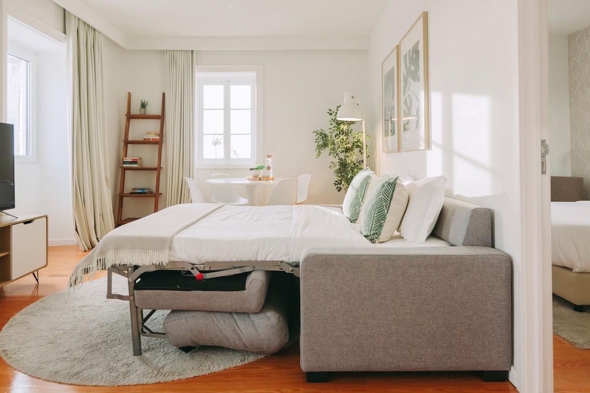 A modern bedroom with a bed, a grey sofa, a small table, a ladder, and a TV.