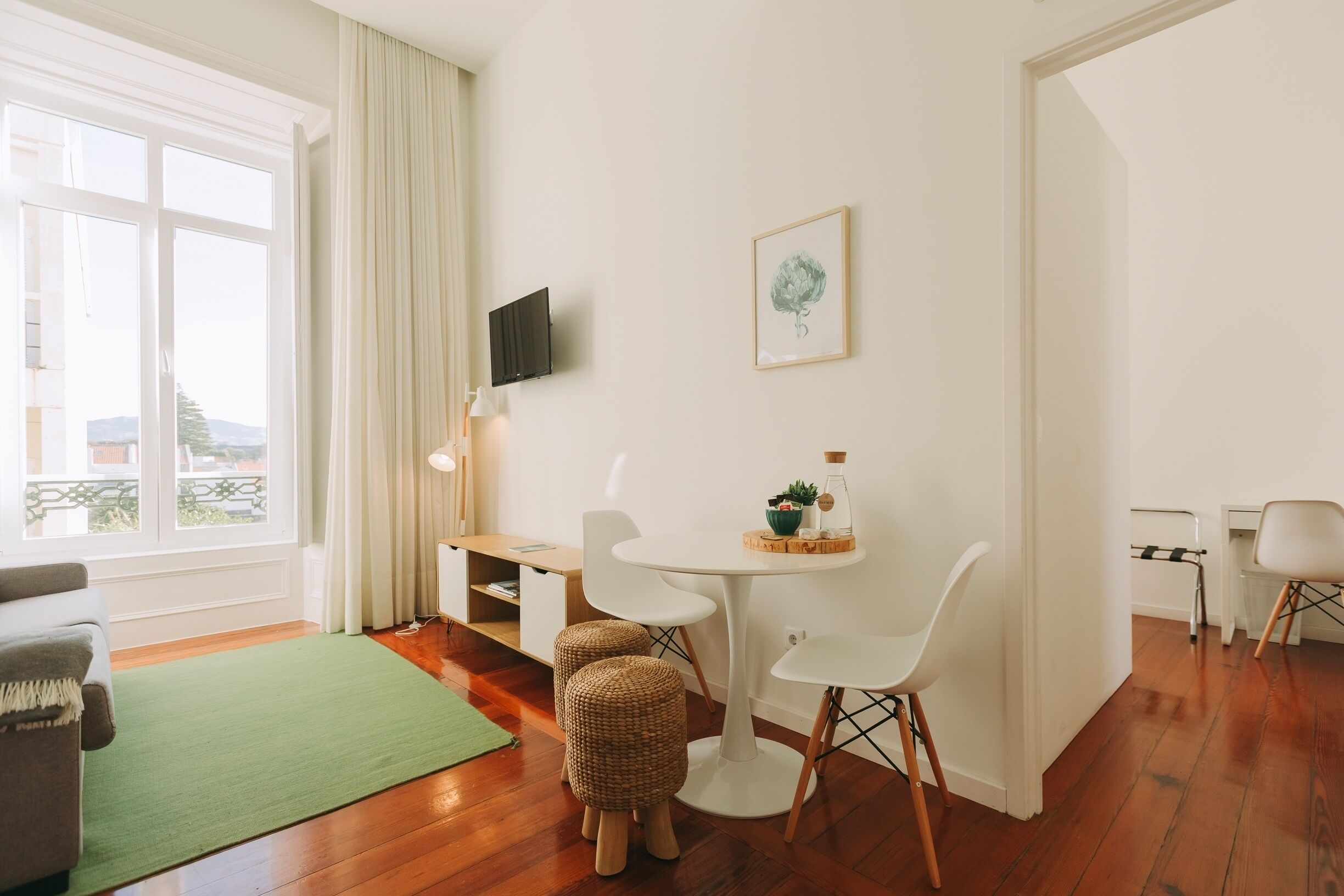 Una sala de estar moderna con un ventanal grande, una mesa de comedor con dos sillas, un televisor montado en la pared y una mesita auxiliar con objetos decorativos.