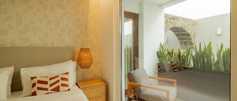A modern bedroom with a bed, bedside table, and a chair by a glass door leading to a balcony with plants.