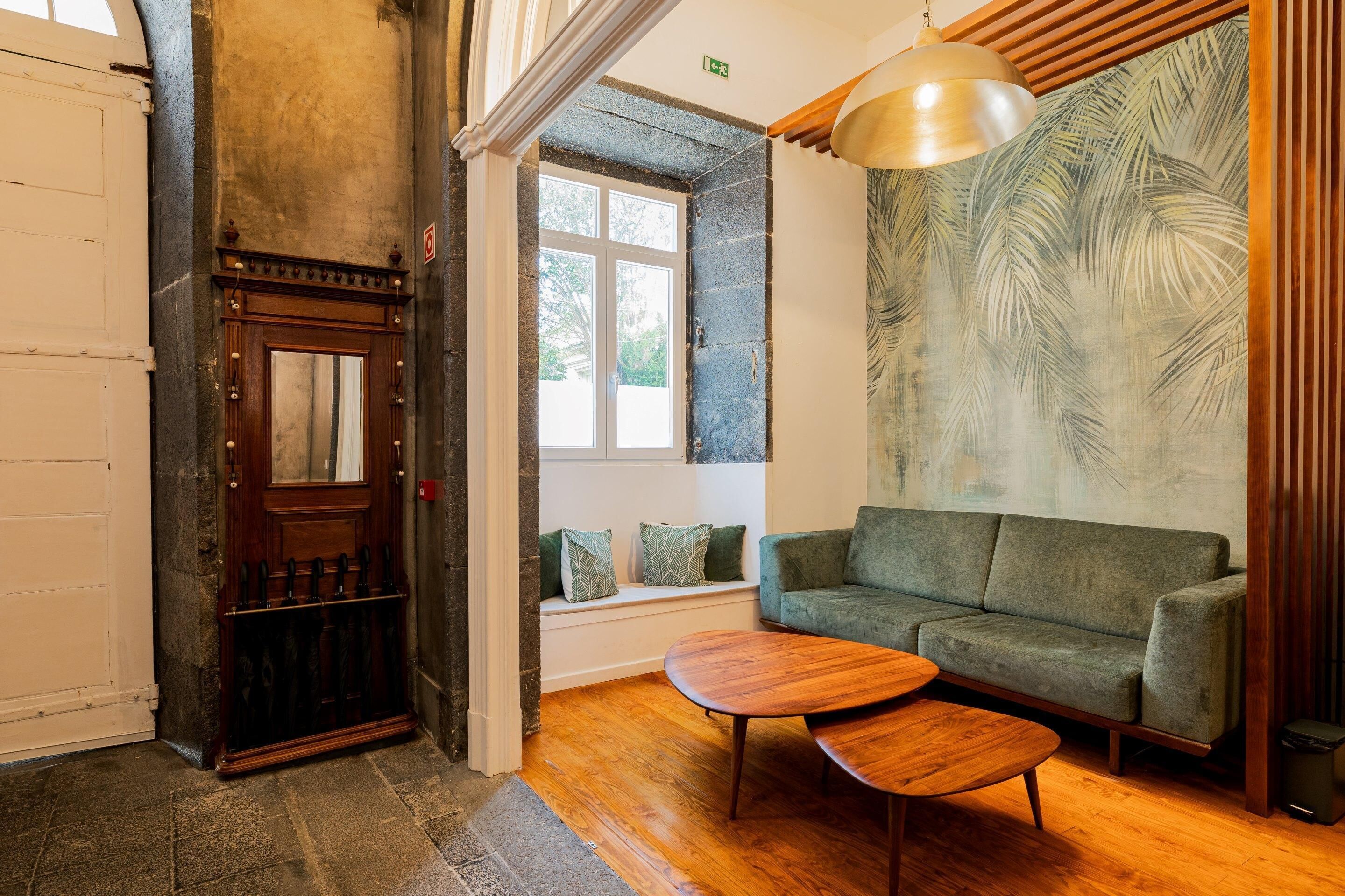 A cozy room with a wooden door, a green sofa, a wooden coffee table, and a large window with a view of trees.