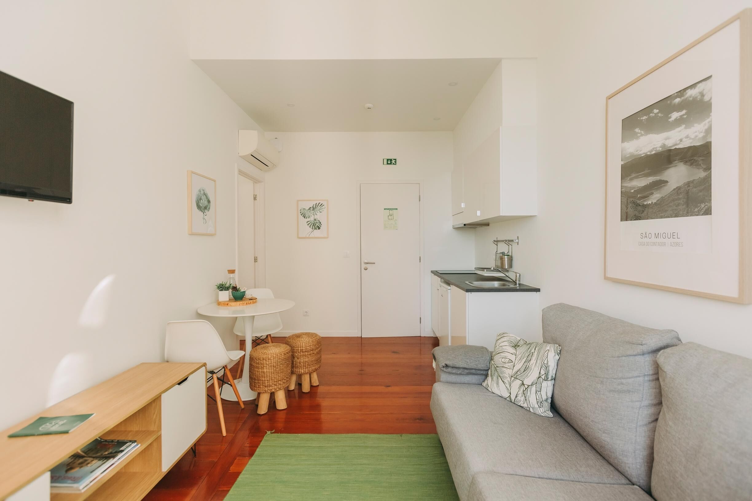Une pièce moderne et bien éclairée, avec un canapé gris, un bureau en bois, une petite table à manger avec des chaises et un coin cuisine.