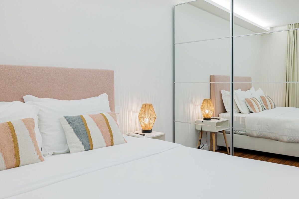 A neatly made bed with striped pillows, a bedside lamp, and a large mirror reflecting the room.