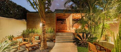 Un patio extérieur moderne, doté de mobilier en bois, d’un chemin en béton et de plantes tropicales.