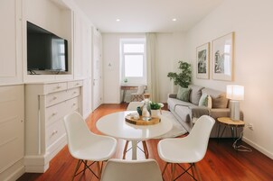 Una sala de estar moderna con televisor, una mesa de comedor con sillas, un sofá, una pequeña mesita de noche con lámpara y una ventana con cortinas.