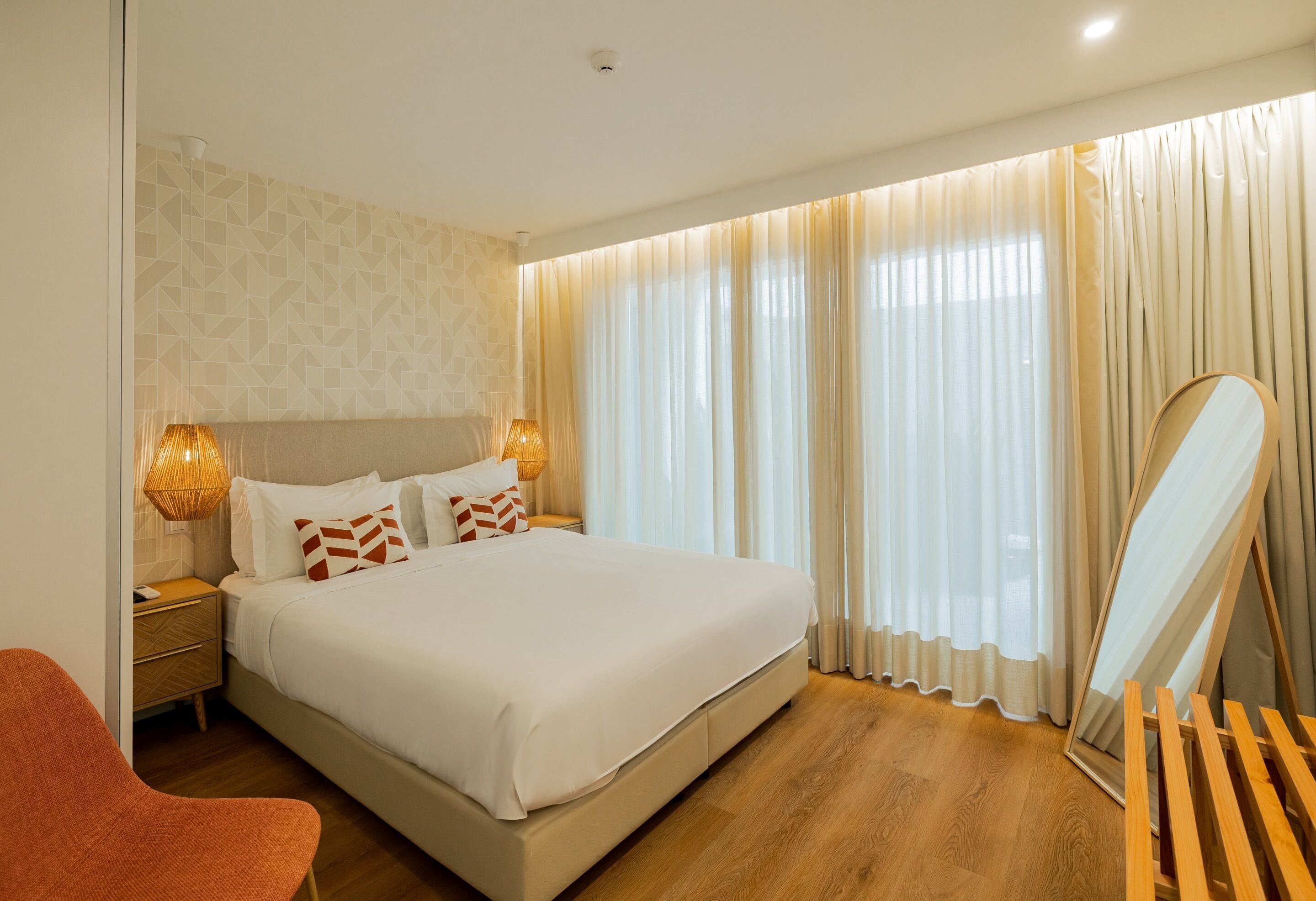 A neatly arranged hotel room with a large bed, a bedside lamp, a mirror, and a wooden chair.