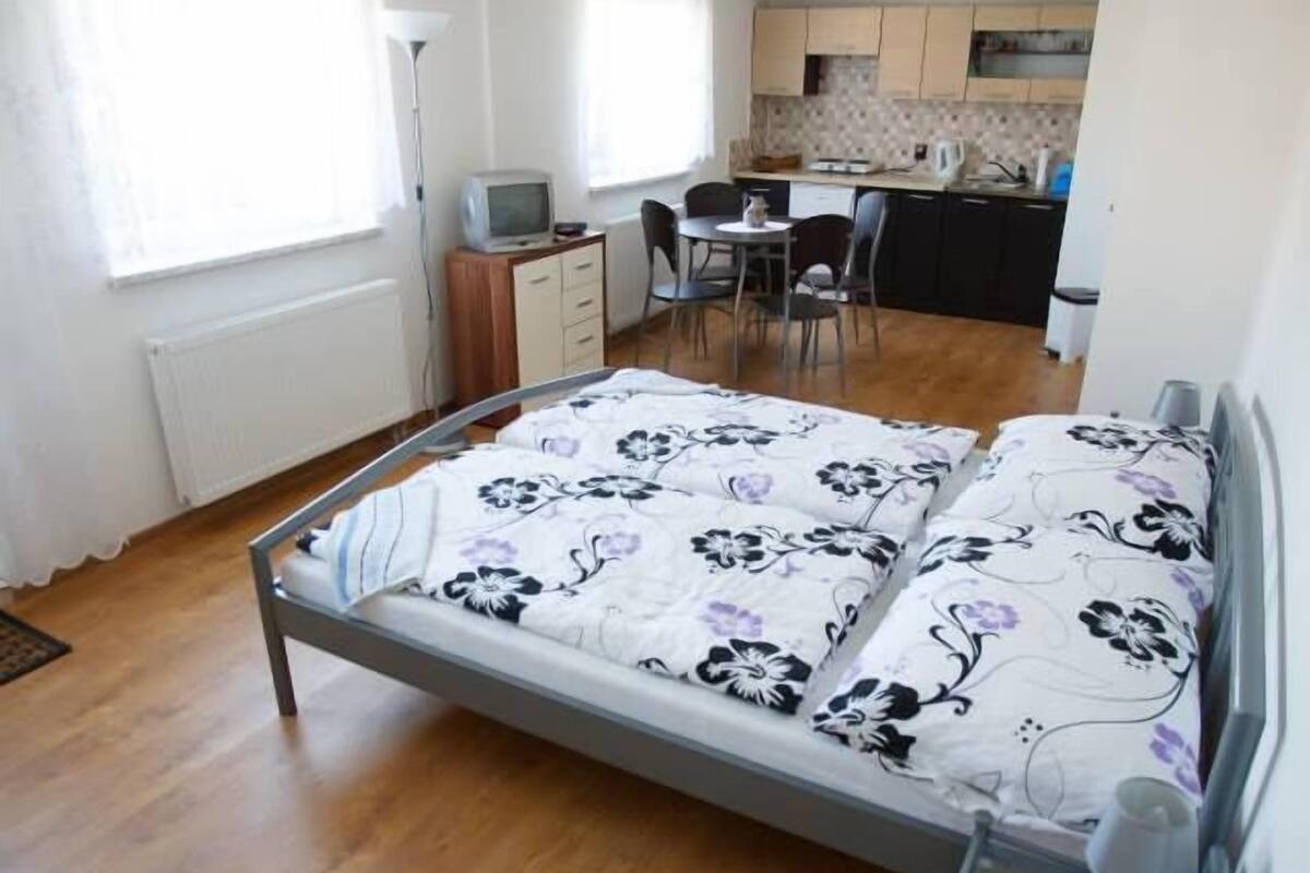 A bedroom with a bed, a desk, and a kitchenette in the background.