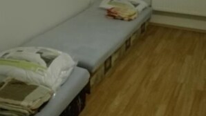 A small, single-bed room with a wooden floor, a window with blinds, and a small table with a newspaper and a bag. - Apartmany Halex 4 (Bojná)