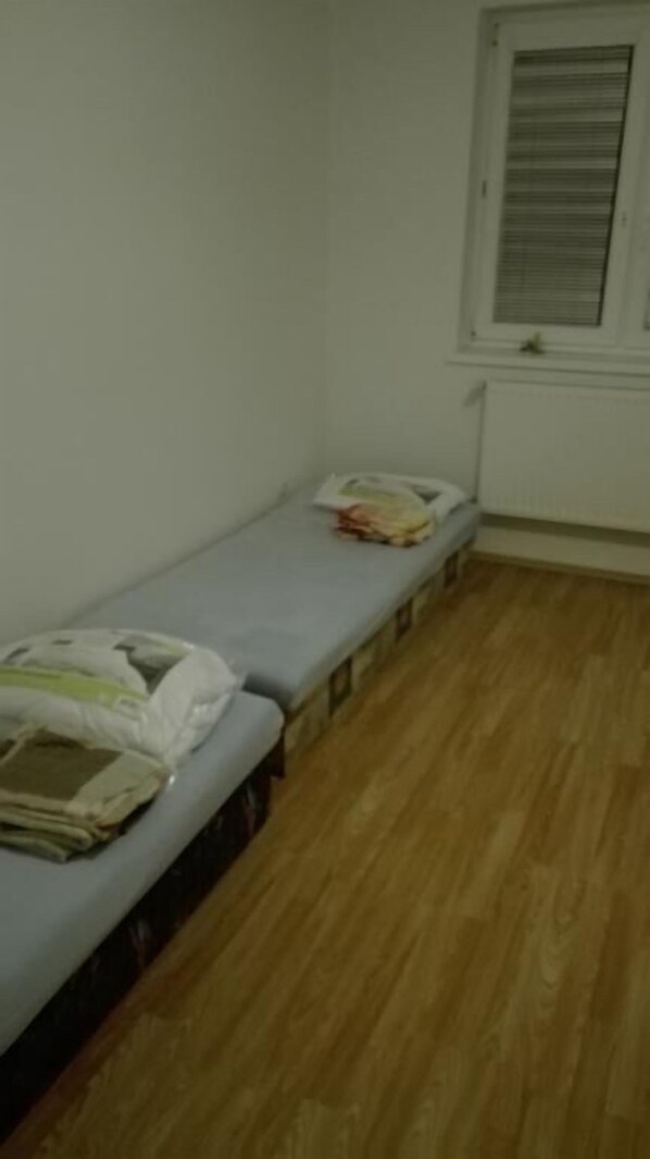 A small, single-bed room with a wooden floor, a window with blinds, and a small table with a newspaper and a bag. - Apartmany Halex 4 (Bojná)