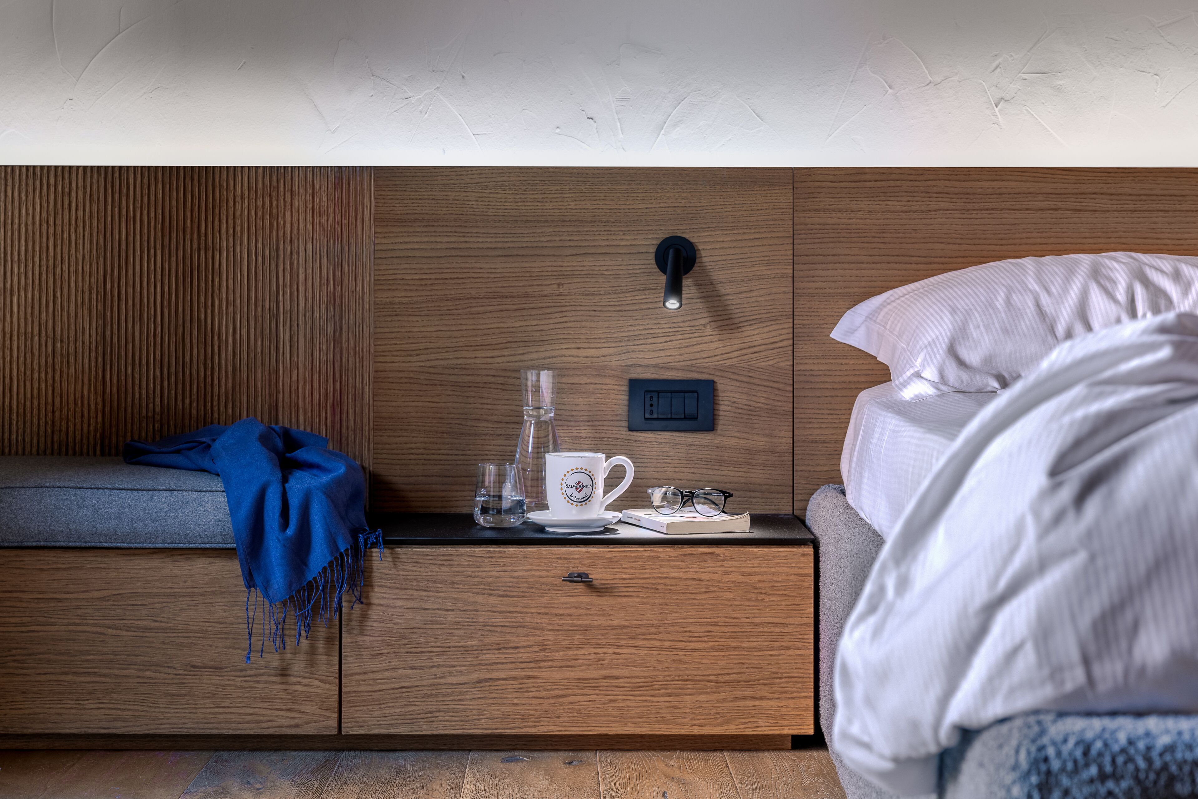 A bedroom with a wooden headboard, a bedside table with a glass, a white mug, and a book.