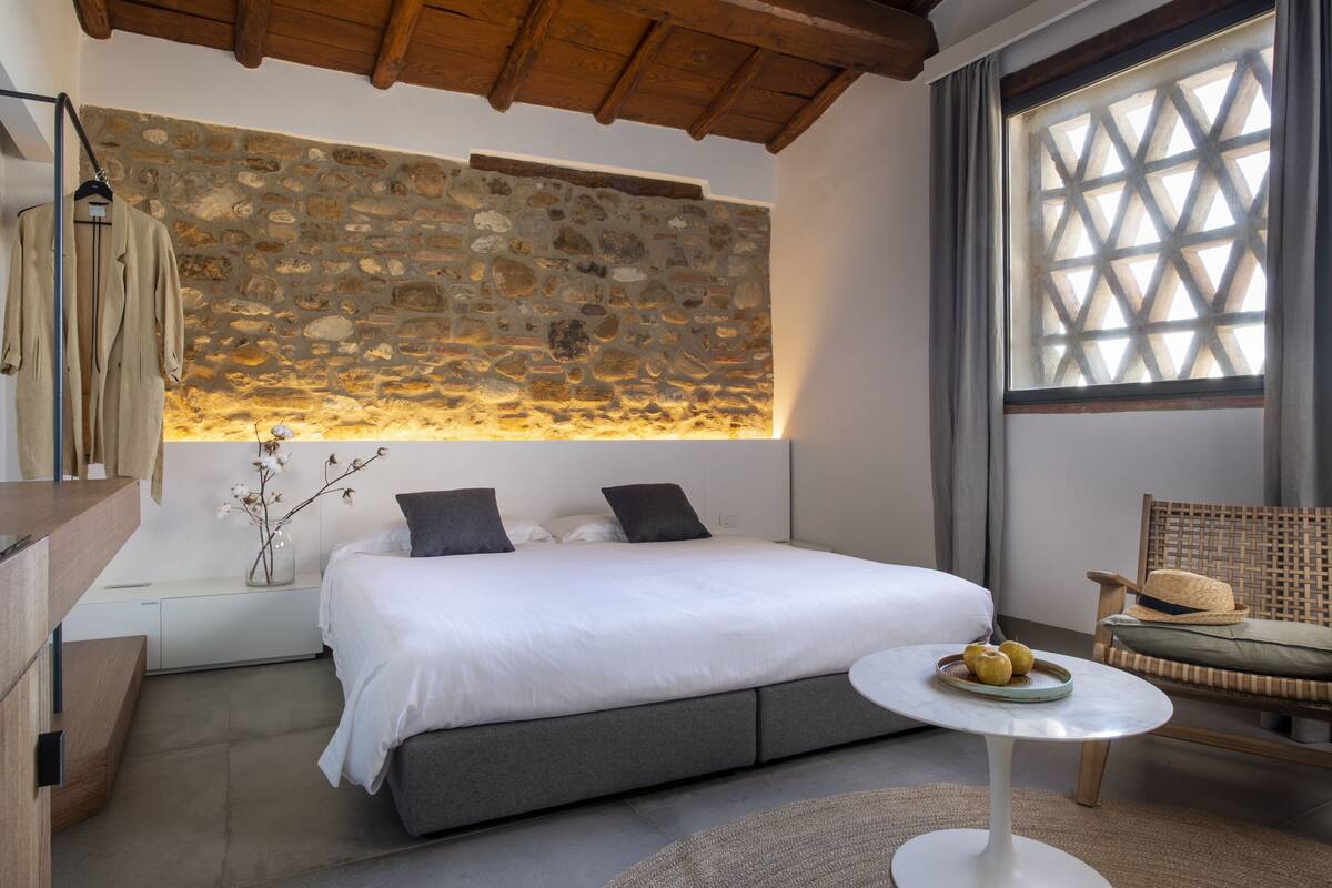 A bedroom with a stone wall, a bed with gray bedding, a round white table with fruit, and a wicker chair.