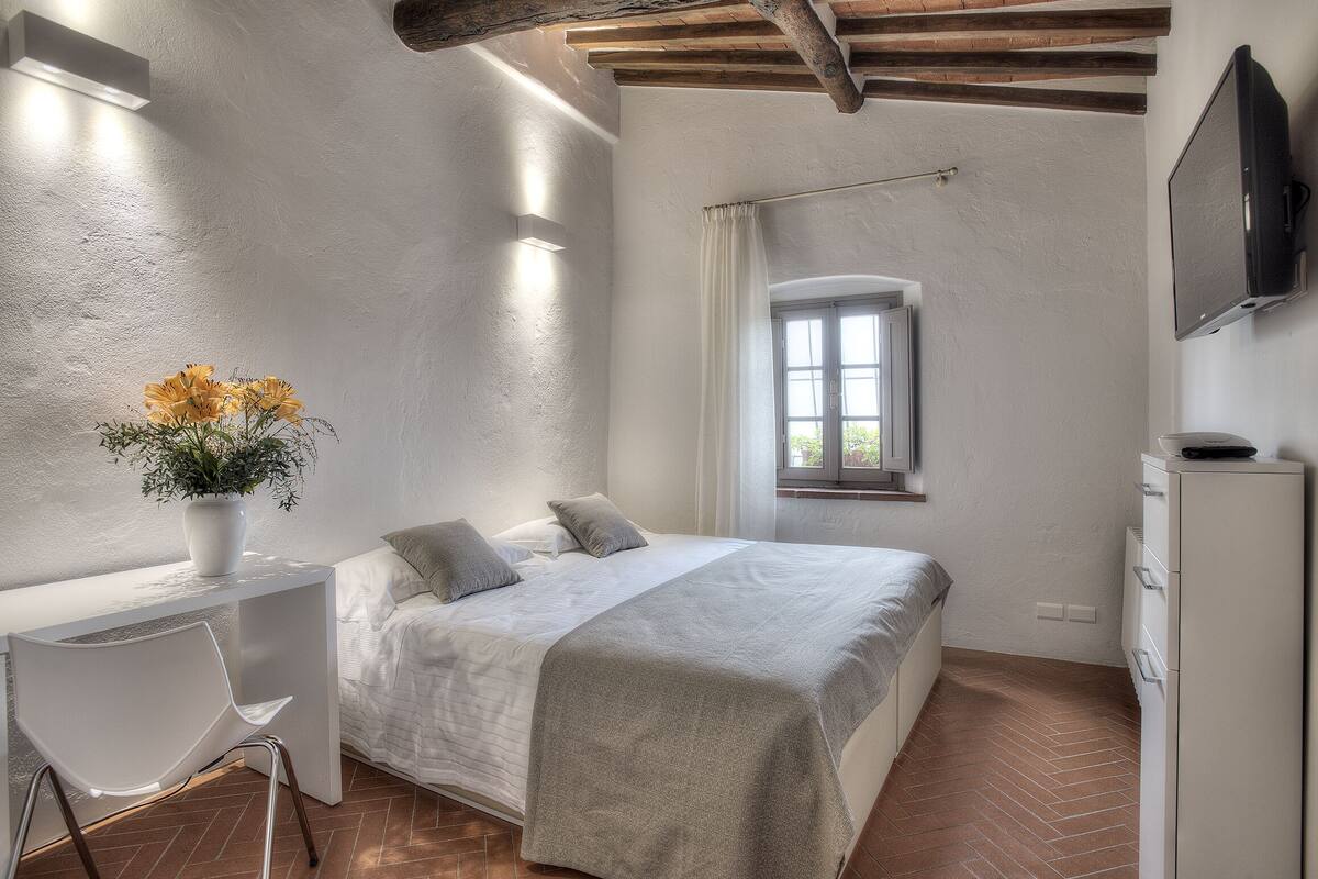 A bedroom with a bed, bedside table, chair, TV, and a window with curtains.