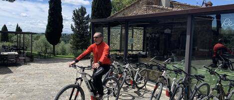 Un hombre pedaleando en bicicleta en un espacio al aire libre con suelo de piedra, rodeado de bicicletas, un área de estar cubierta y un edificio de piedra con cerramiento de vidrio.