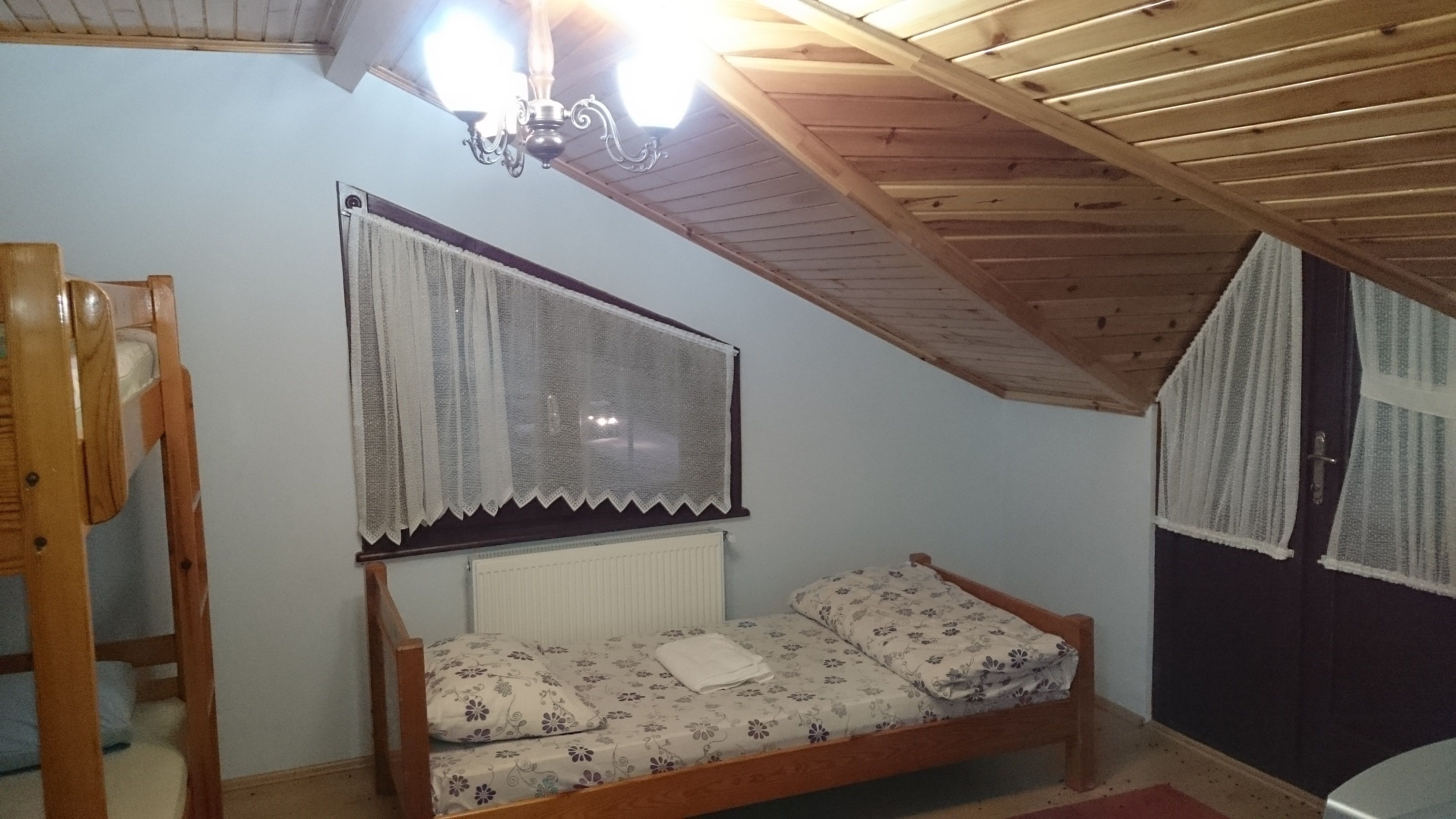 A small, cozy room with a wooden ceiling, a single bed with floral bedding, a radiator, and a window with lace curtains.