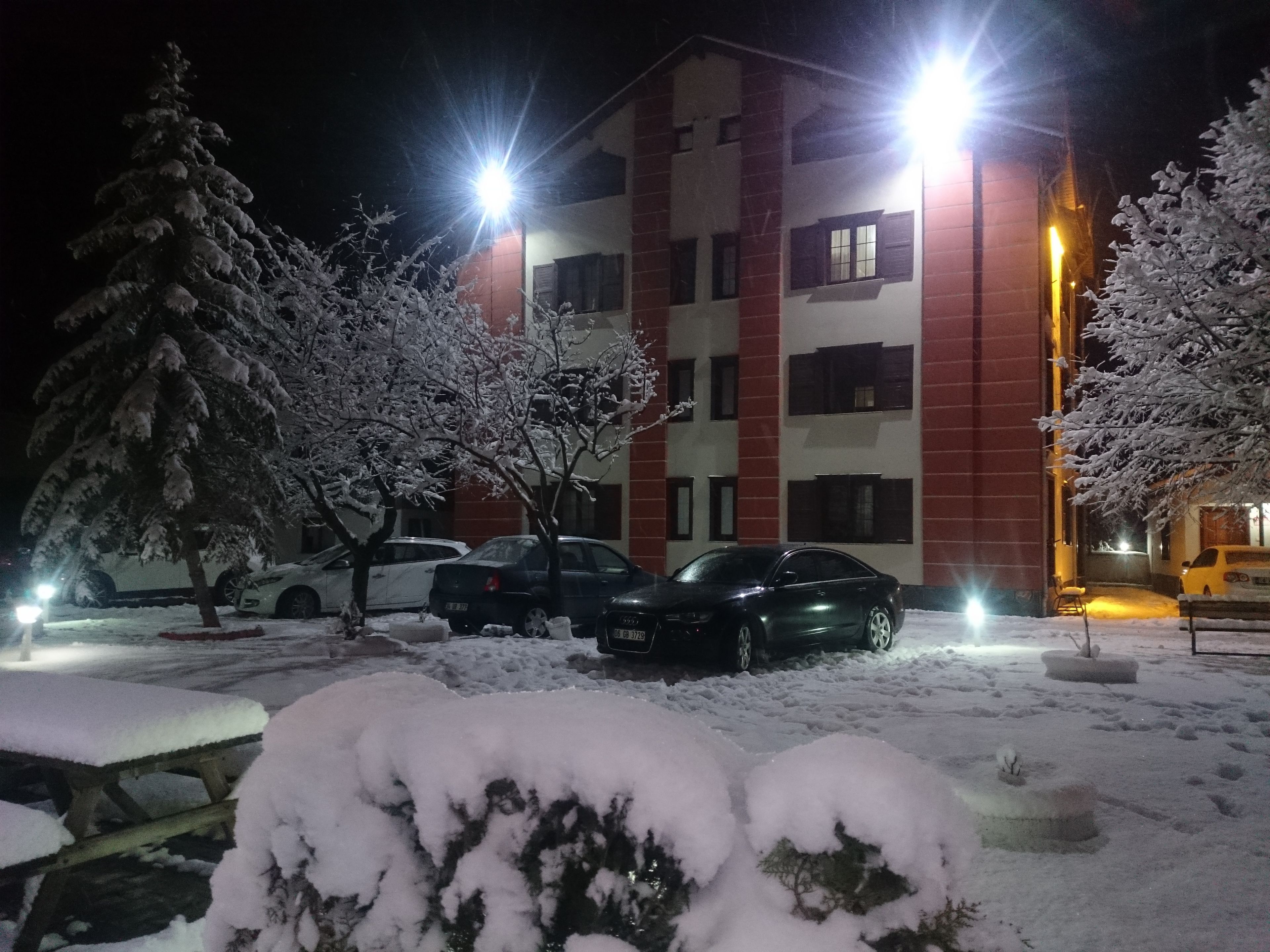 Un edificio de varios pisos con fachada roja y blanca, varios coches aparcados y árboles cubiertos de nieve.