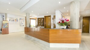 A reception desk with a wooden finish, a white base, and a large pillar to the right. There is a bulletin board with information and a flower arrangement on the desk. - Apartamentos Massol (Pollenca)