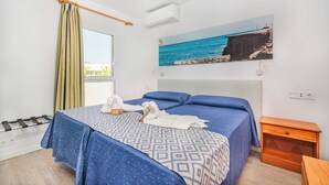 A hotel room with a bed, a painting of a coastal scene, a window with curtains, and a wooden nightstand. - Apartamentos Massol (Pollenca)