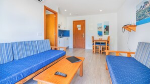 A room with a sofa, a dining table with chairs, a small kitchen area, and a door leading to another room. - Apartamentos Massol (Pollenca)