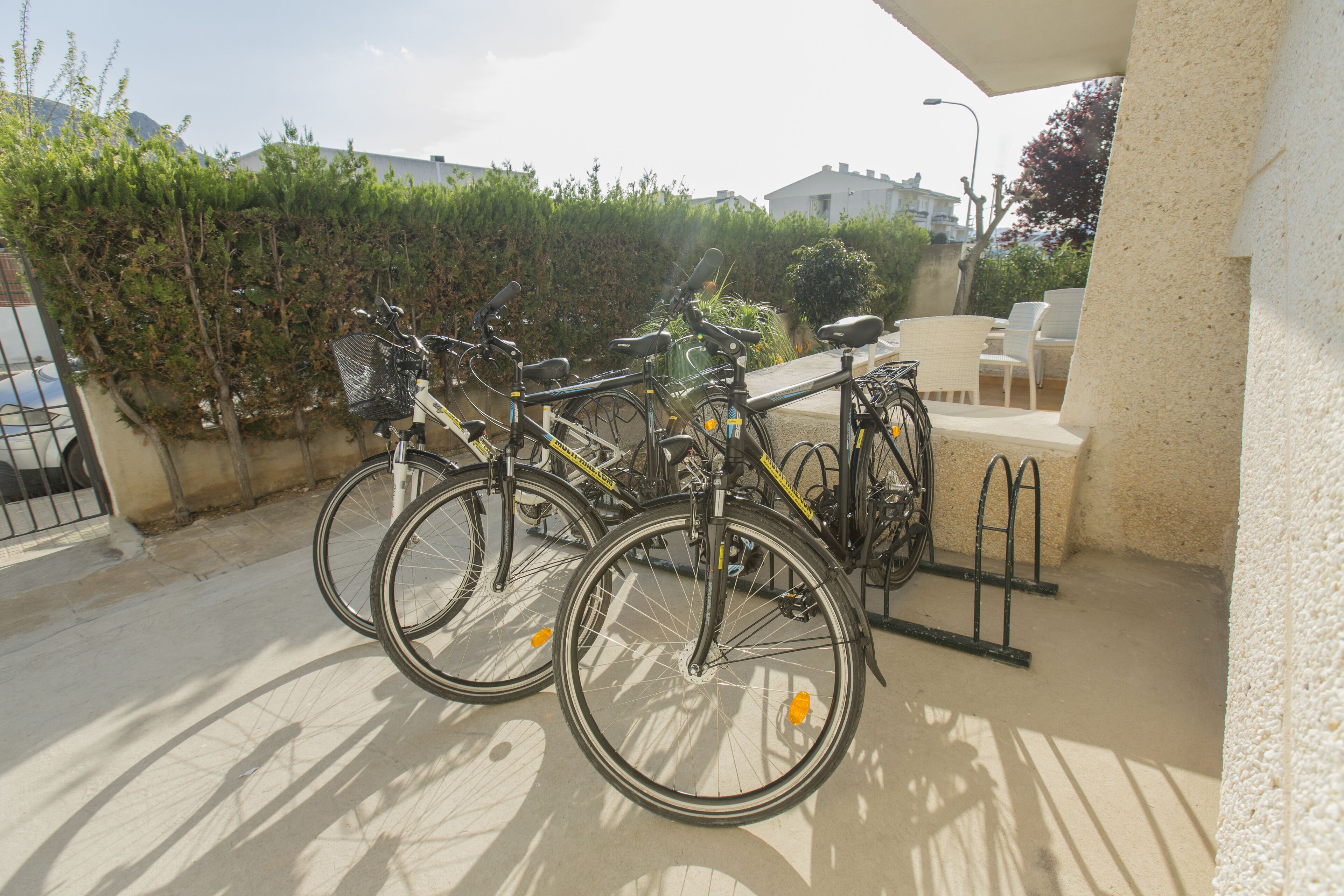 Bicycles parked under a covered area with a white wall and a hedge in the background.