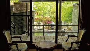 Habitación con un ventanal grande que da a una terraza de madera y jardín. Hay dos sillones tapizados y una pequeña mesa redonda.
