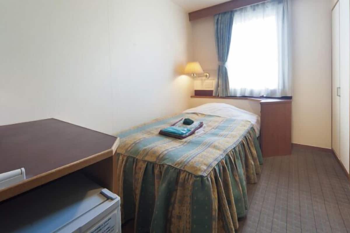 Ett hotellrum med en säng, ett skrivbord, en lampa och ett fönster med gardiner.