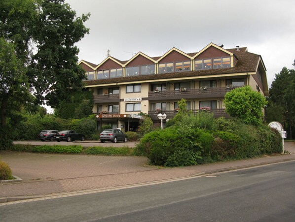 A multi-story hotel with a row of balconies, a parking area, and lush greenery in front.