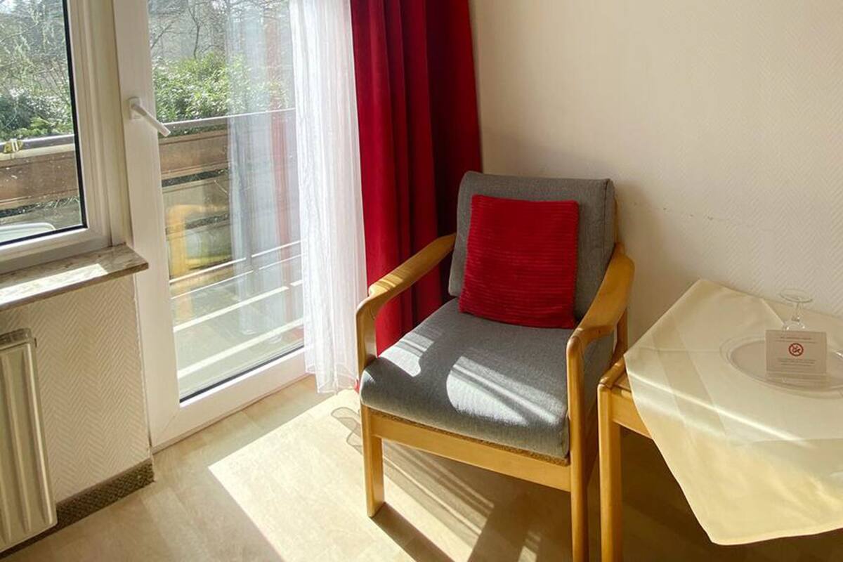 Ein Zimmer mit einem Sessel, einem Tisch mit einem Glas und einem Fenster mit Vorhängen.