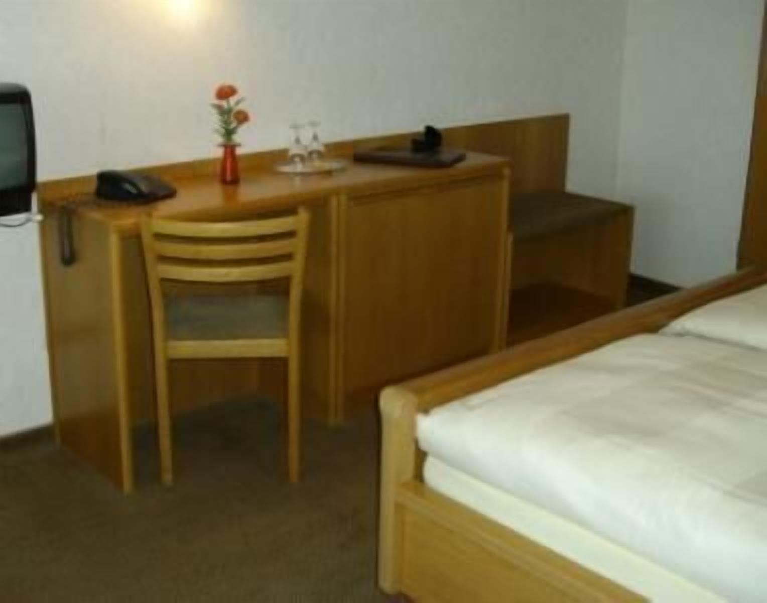 Une chambre d’hôtel de taille modeste, comprenant un bureau en bois, une chaise, un lit et une petite table de chevet.