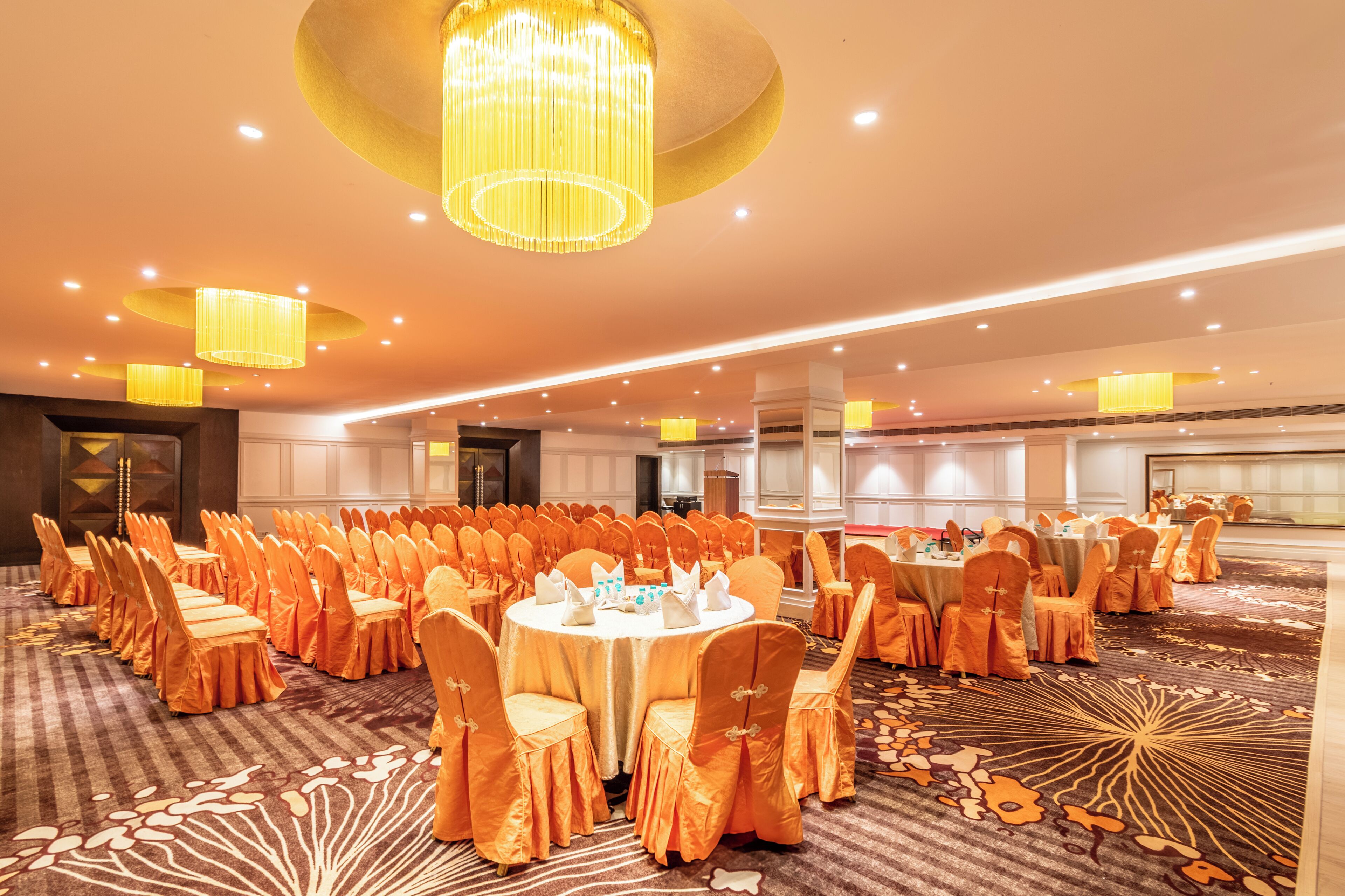 A banquet hall with round tables, orange chairs, and a carpet with a floral pattern.