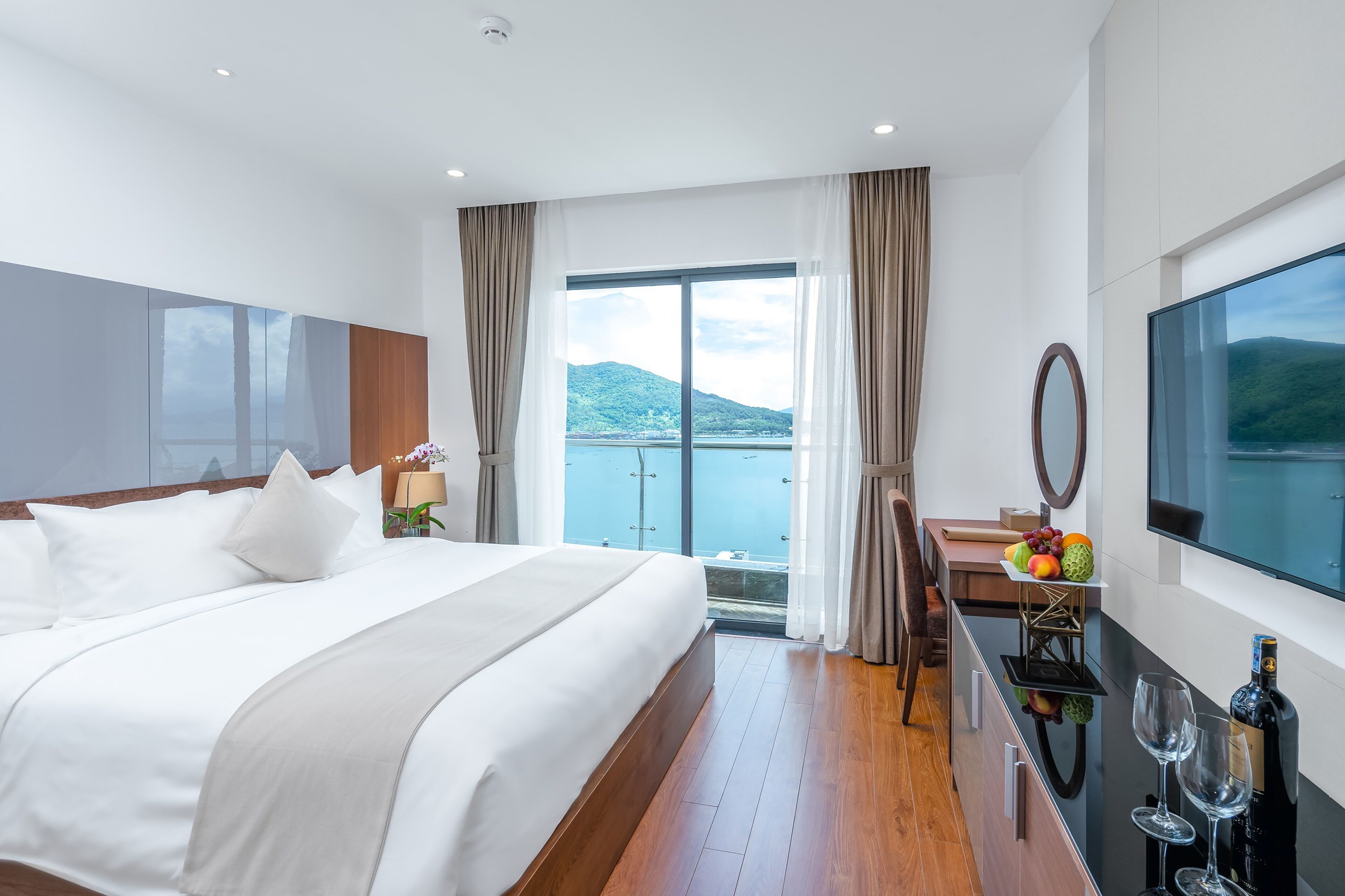 Una habitación de hotel moderna con una cama grande, un escritorio con espejo, un televisor y un balcón con vistas panorámicas.