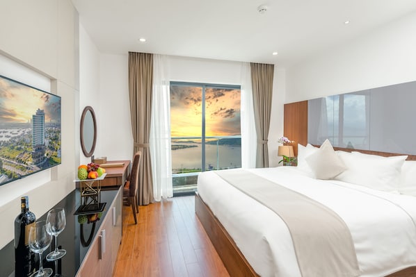 A modern hotel room with a large bed, a desk with a mirror, a view of the sunset, and a cityscape painting. - Alan Sea Da Nang (Da Nang)