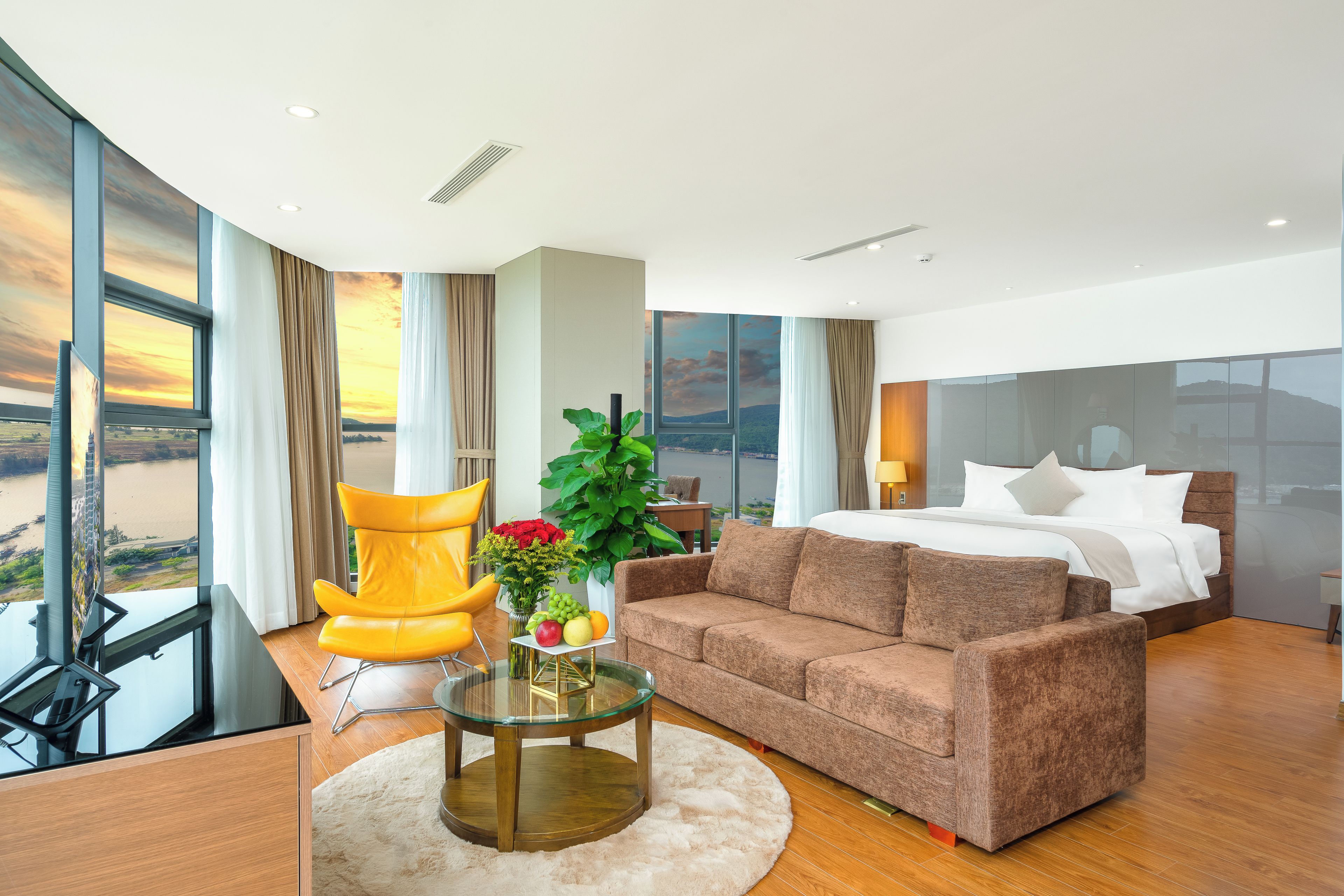 A modern hotel room with a large bed, a sofa, a round glass table, and a view of a sunset over a body of water.