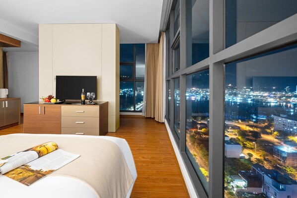 Een moderne hotelkamer met een groot bed, een televisie op een houten kast en uitzicht op de stad door grote ramen.