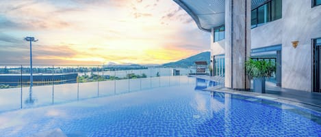 Uma piscina infinita moderna com vista clara do pôr do sol sobre a água, cercada por um edifício contemporâneo com amplas janelas e telhado curvo.