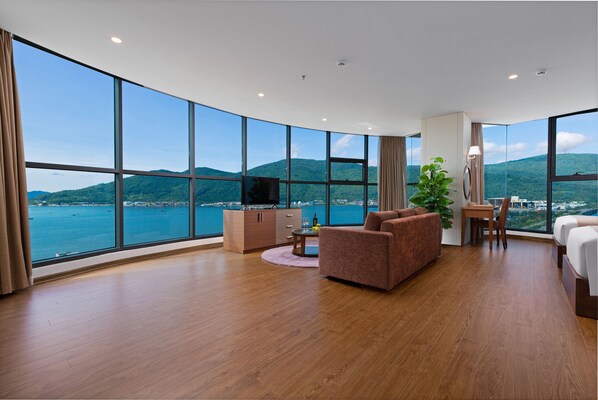 Amplia habitación con ventanas grandes que ofrecen vistas al agua y colinas. Cuenta con un sofá, una pequeña mesa y un televisor.