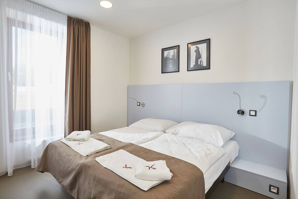 A hotel room with a large bed, two pillows, a headboard, a bedside table, a nightstand, a window with sheer curtains, and two framed pictures on the wall.
