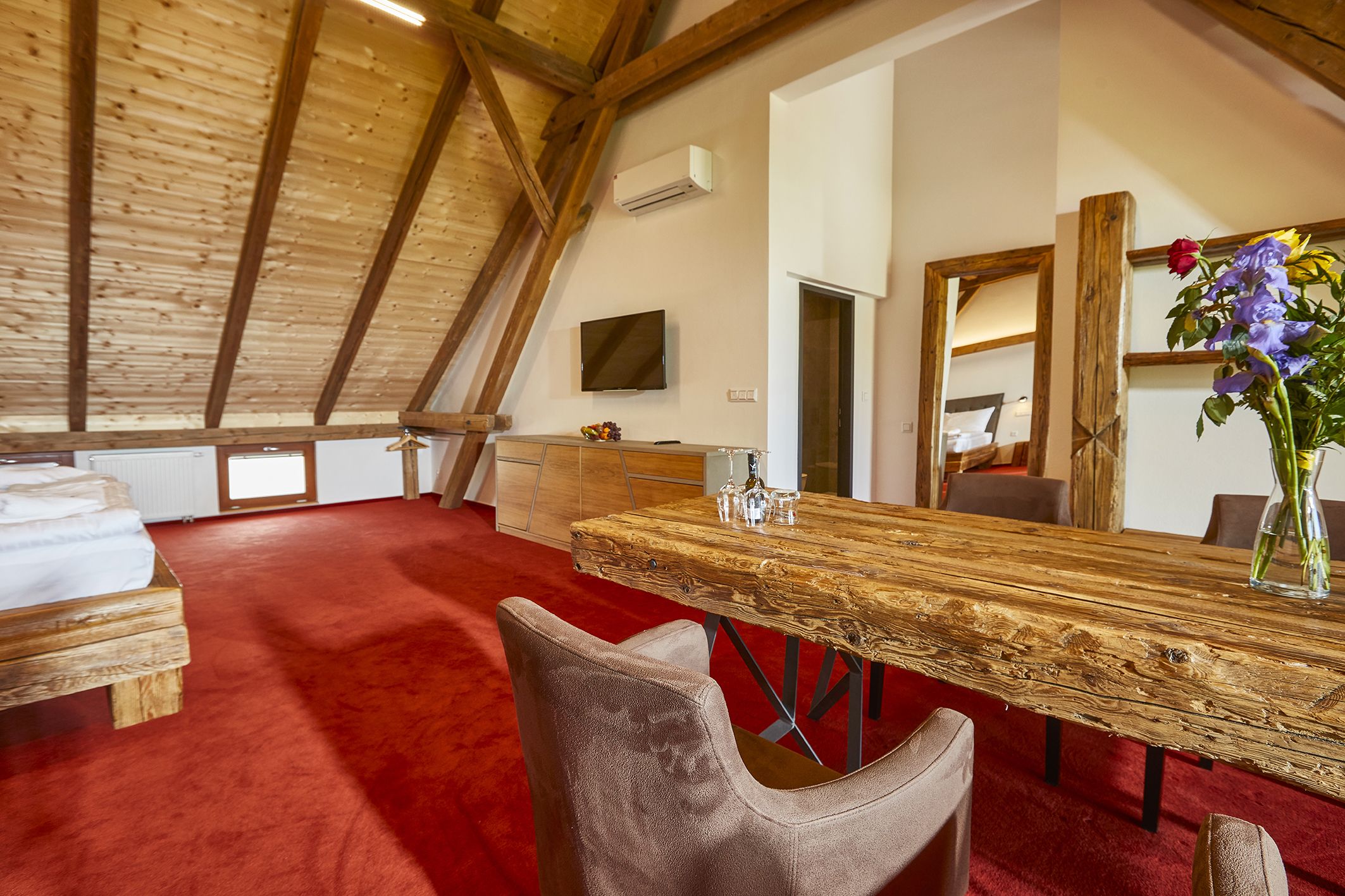 A spacious room with a high ceiling, a wooden ceiling, a red carpet, a wooden dining table, and a mirror.