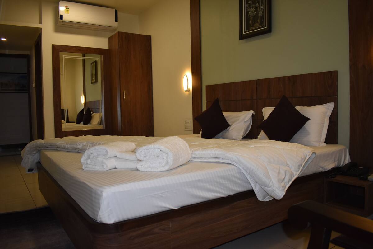 A neatly made bed with white linens and rolled towels, a wooden headboard, a bedside table with a phone, and a wall-mounted air conditioner.