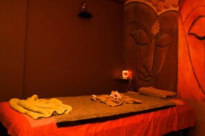 A massage room with a bed, towels, a flower, and a wall with a face design. - Hotel Shyam Palace (Bhuj)