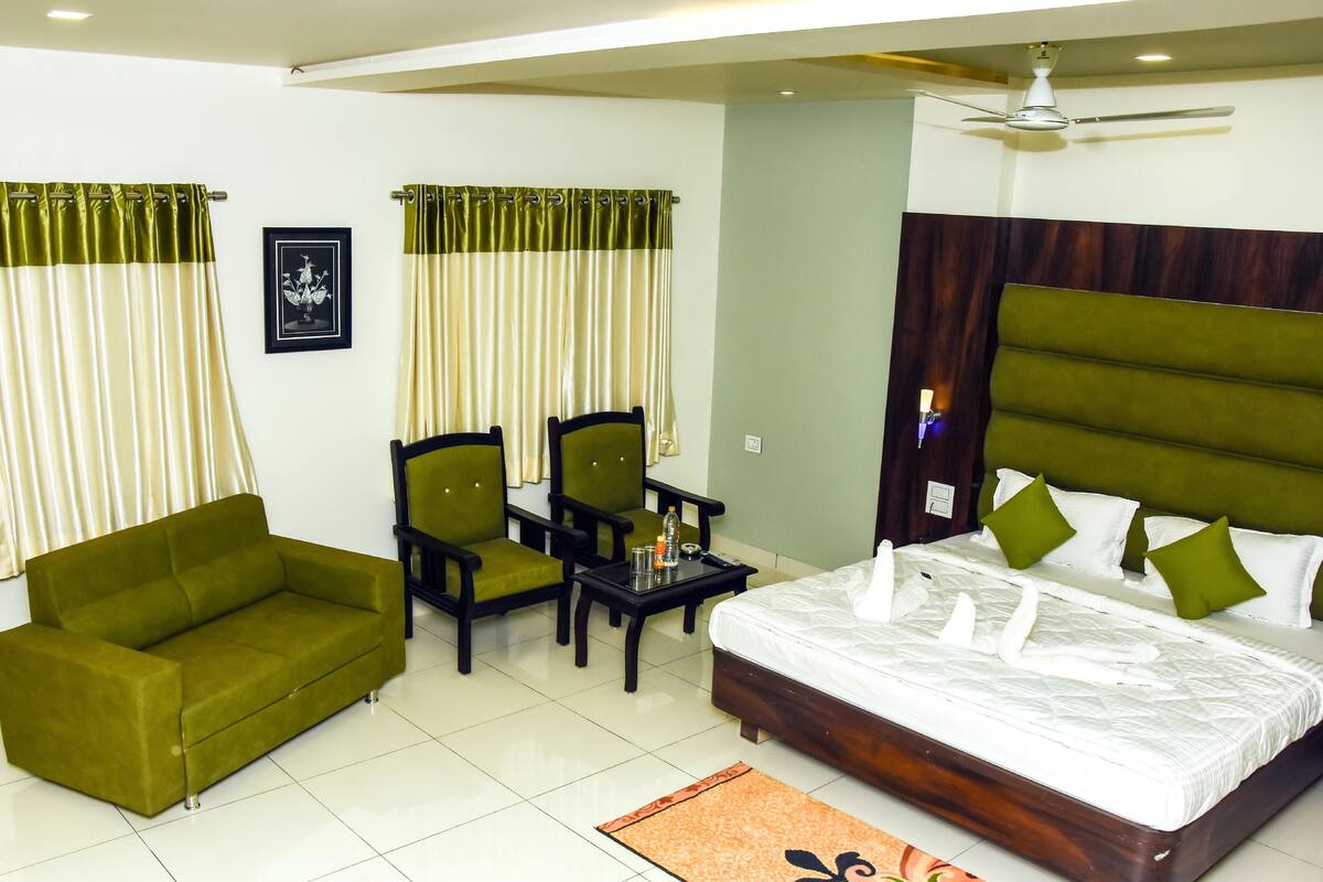 A hotel room with a green sofa, two chairs, a small table, a bed with white bedding and green pillows, and a ceiling fan.