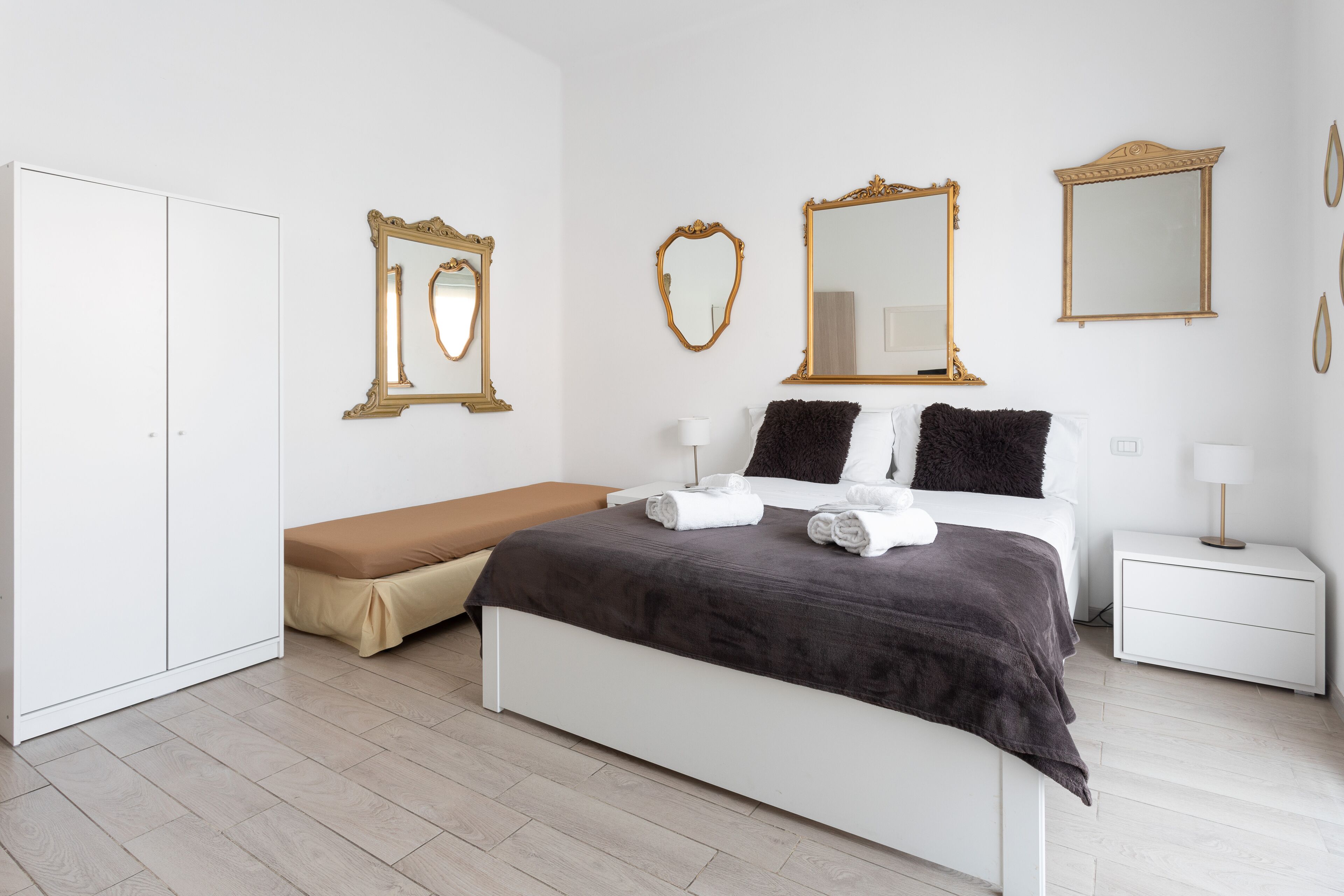 Un dormitorio con una cama, un banco, una mesita de noche y tres espejos.