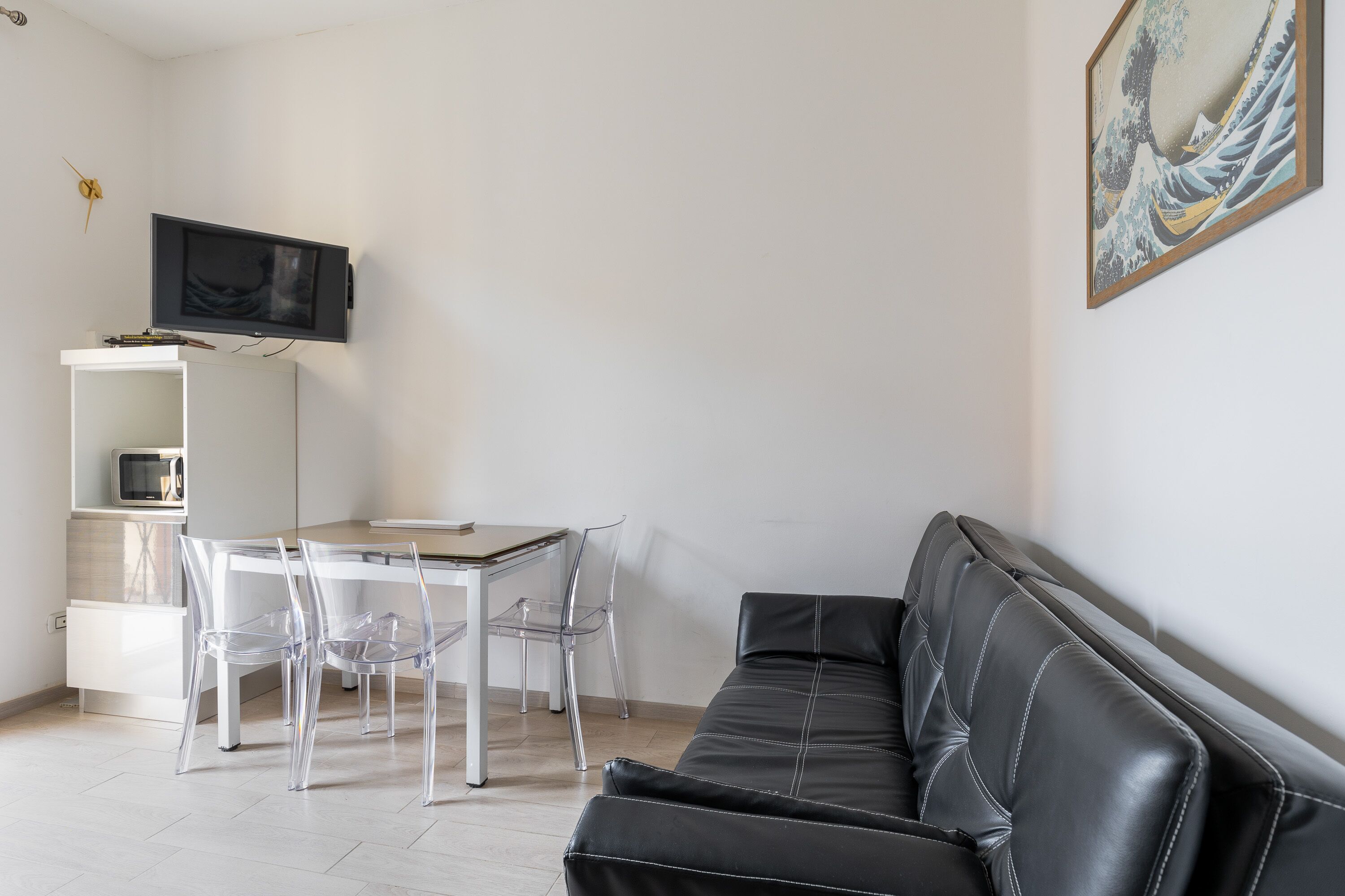 A modern living room with a black leather sofa, a dining table with transparent chairs, a flat-screen TV mounted on the wall, and a framed artwork above the sofa.