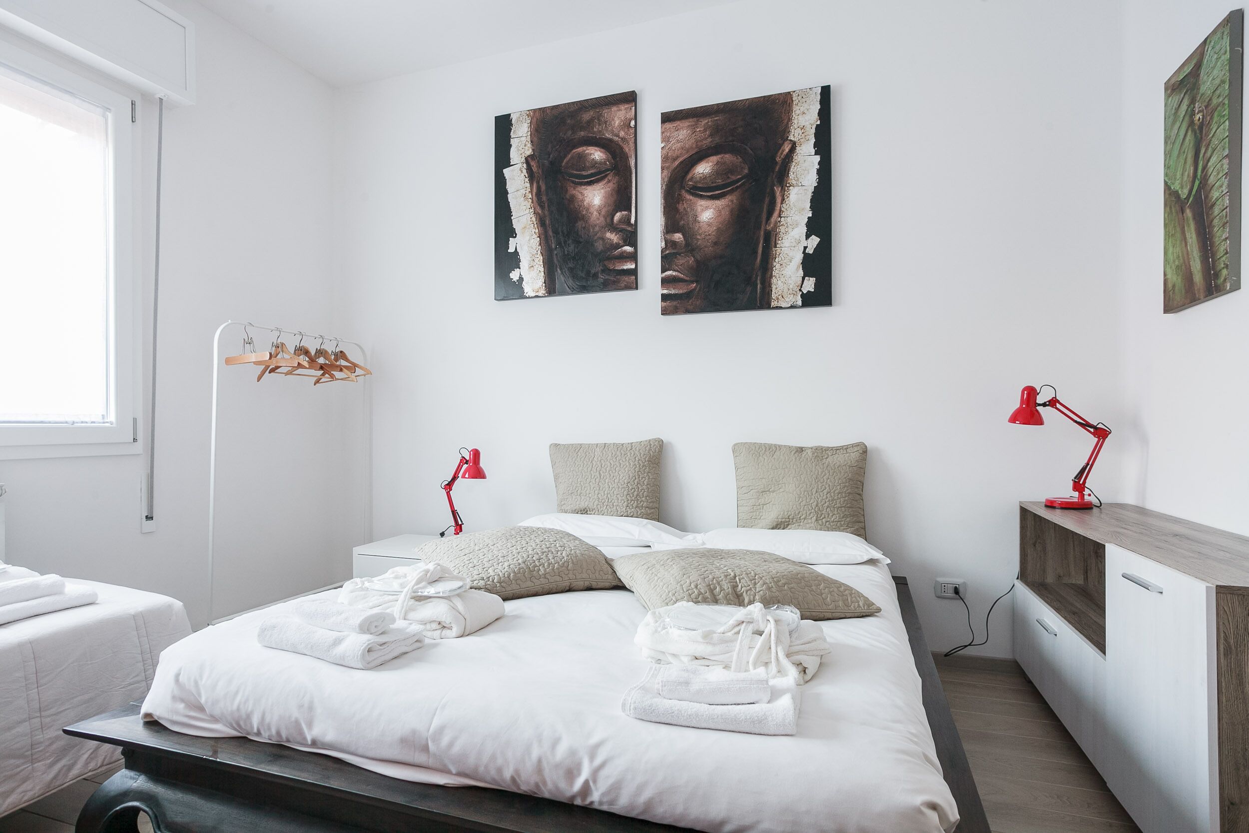 A modern hotel room with a neatly made bed, two wall-mounted artworks, a red desk lamp, and a wooden side table.