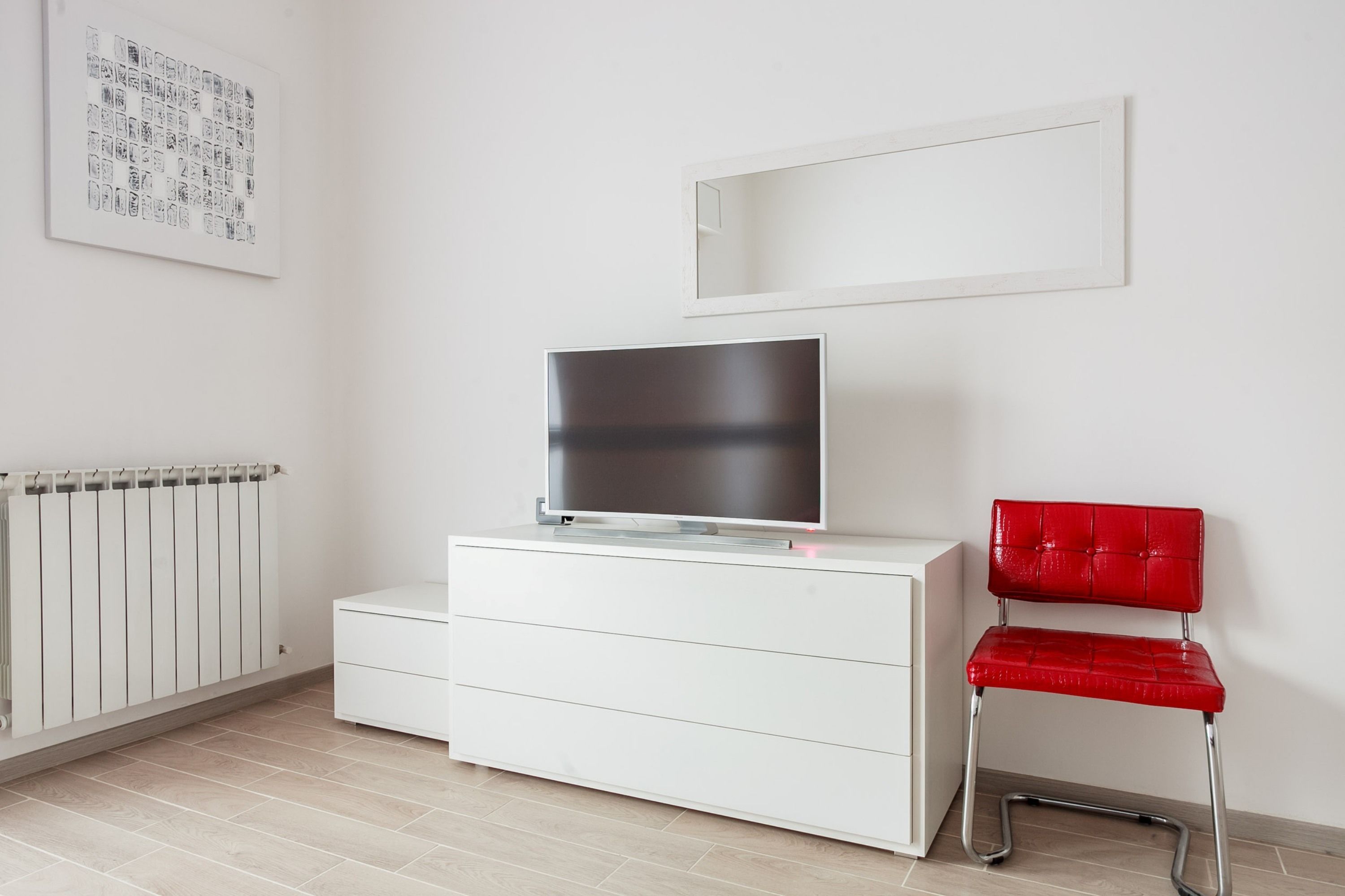 A room with a white dresser, a red chair, a television on the dresser, a radiator, and a framed picture on the wall.