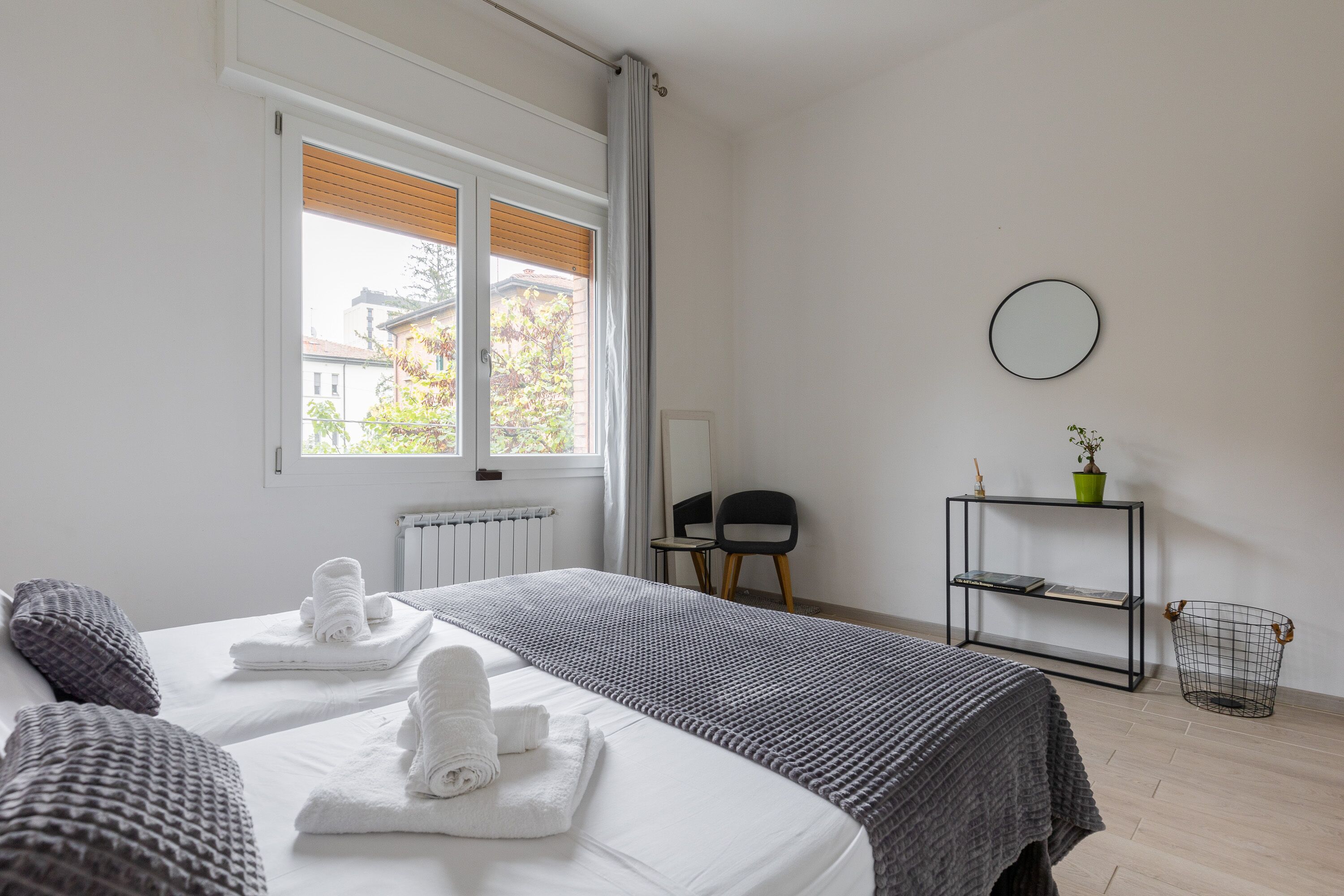 Ein Hotelzimmer mit zwei Betten, einem Fenster mit Rollo, einem Heizkörper, einem kleinen Tisch mit Stühlen, einem Regal mit Pflanze und einem runden Spiegel an der Wand.