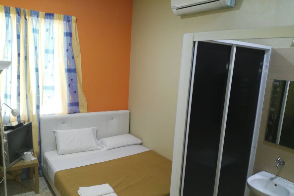 Une chambre d’hôtel comprenant un lit, une table de chevet avec une lampe, une fenêtre avec des rideaux, un appareil de climatisation et une salle de bain avec un lavabo et un miroir.