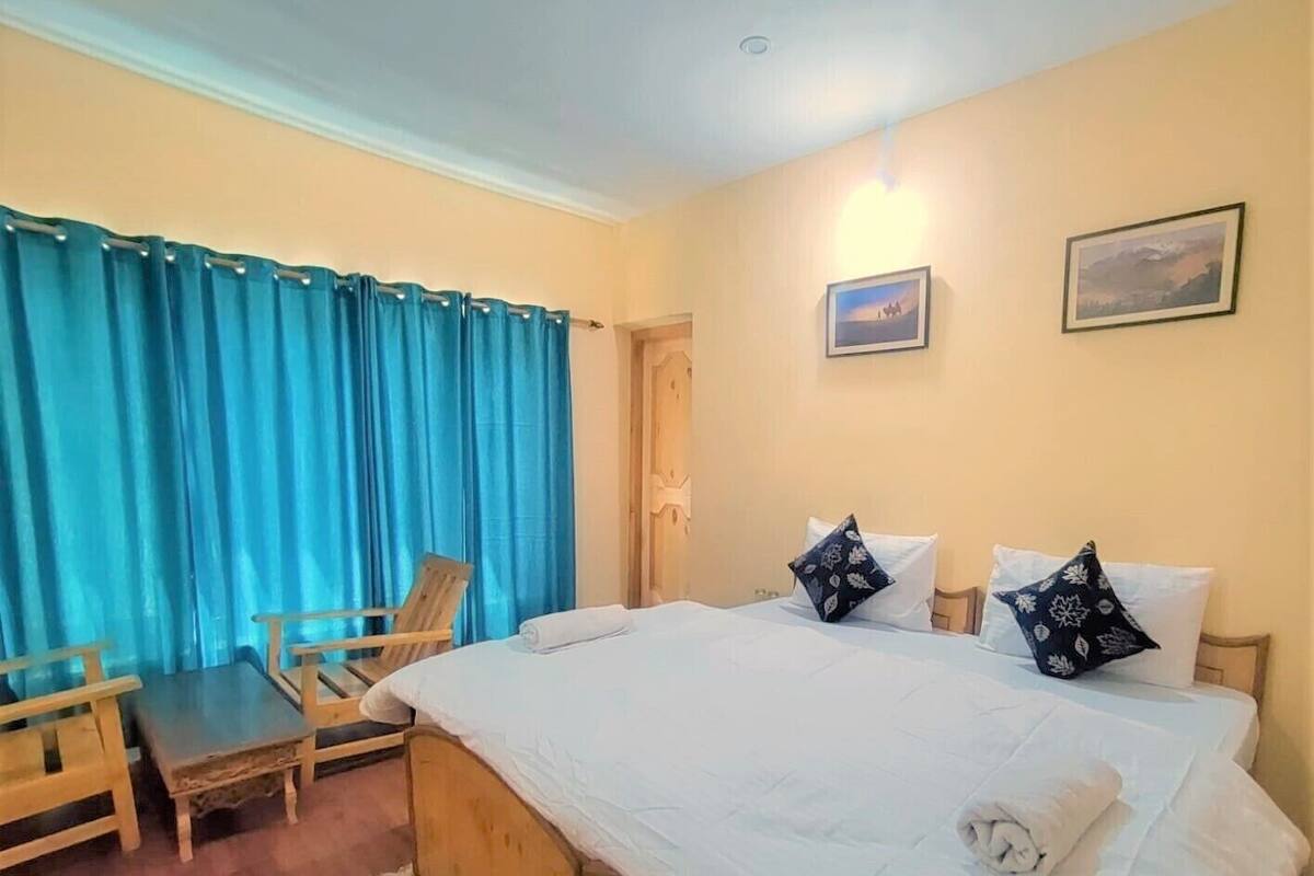 Una habitación de hotel con una cama grande, dos almohadas, una silla de madera, una cortina azul y una alfombra pequeña.